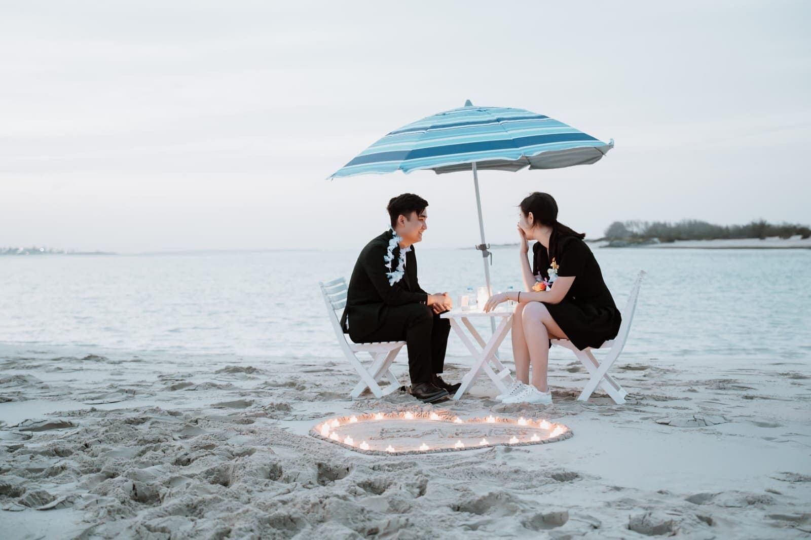 Couple sharing a private table under a beach umbrella beside a heart of candles in the sand